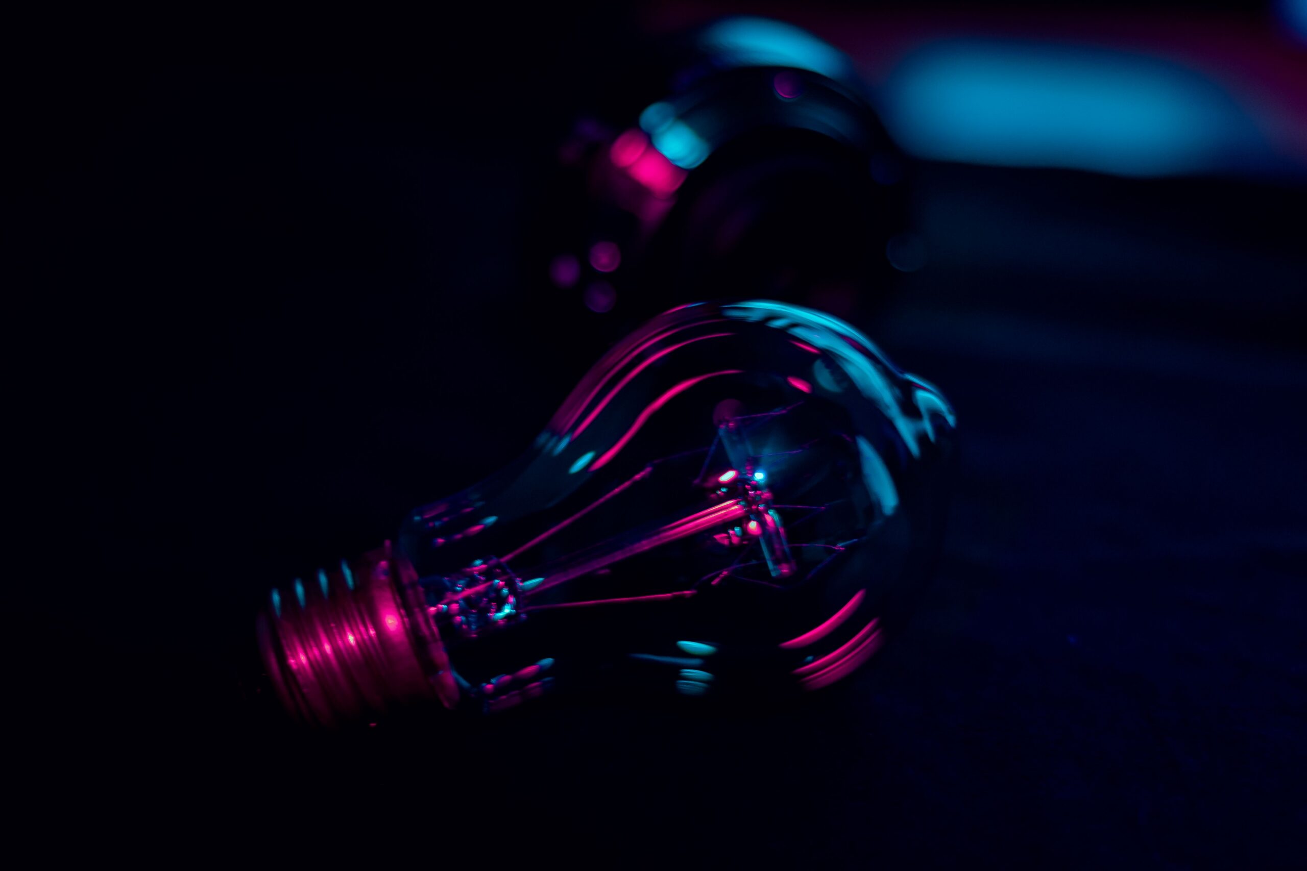 A close-up photograph of a glass light bulb against a dark background, lit with pink and teal neon light that illuminates the internal filament.