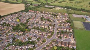 An aerial photograph of a residential neighbourhood in England, showing rows of brick houses with gardens, curved streets, and parked cars. Green fields, sports courts, and open countryside are visible at the edges of the estate.