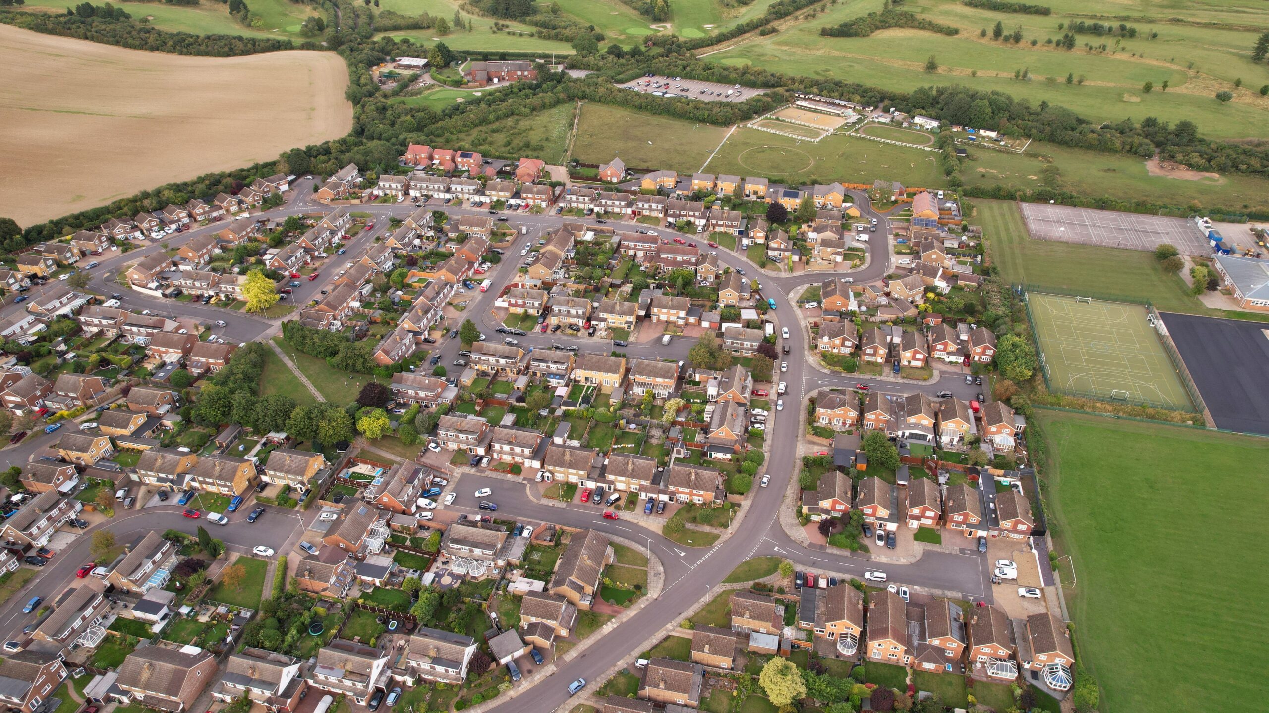 An aerial photograph of a residential neighbourhood in England, showing rows of brick houses with gardens, curved streets, and parked cars. Green fields, sports courts, and open countryside are visible at the edges of the estate.